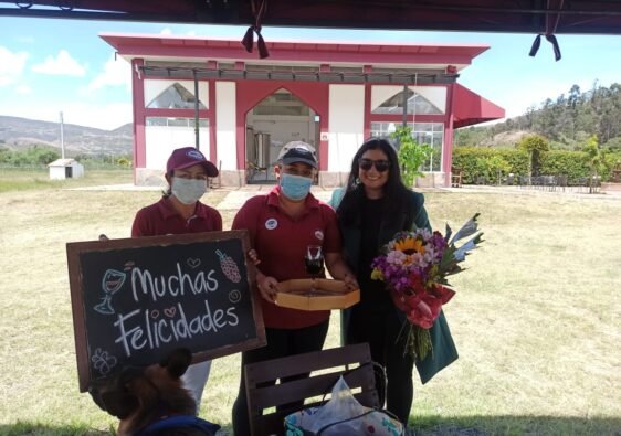 Celebración de cumpleaños por dos trabajadoras del Viñedo Umaña Dajud, un cartel y una copa de vino. Diana posa al lado de ellas con un ramo de flores