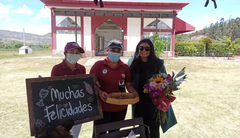 Celebración de cumpleaños por dos trabajadoras del Viñedo Umaña Dajud, un cartel y una copa de vino. Diana posa al lado de ellas con un ramo de flores