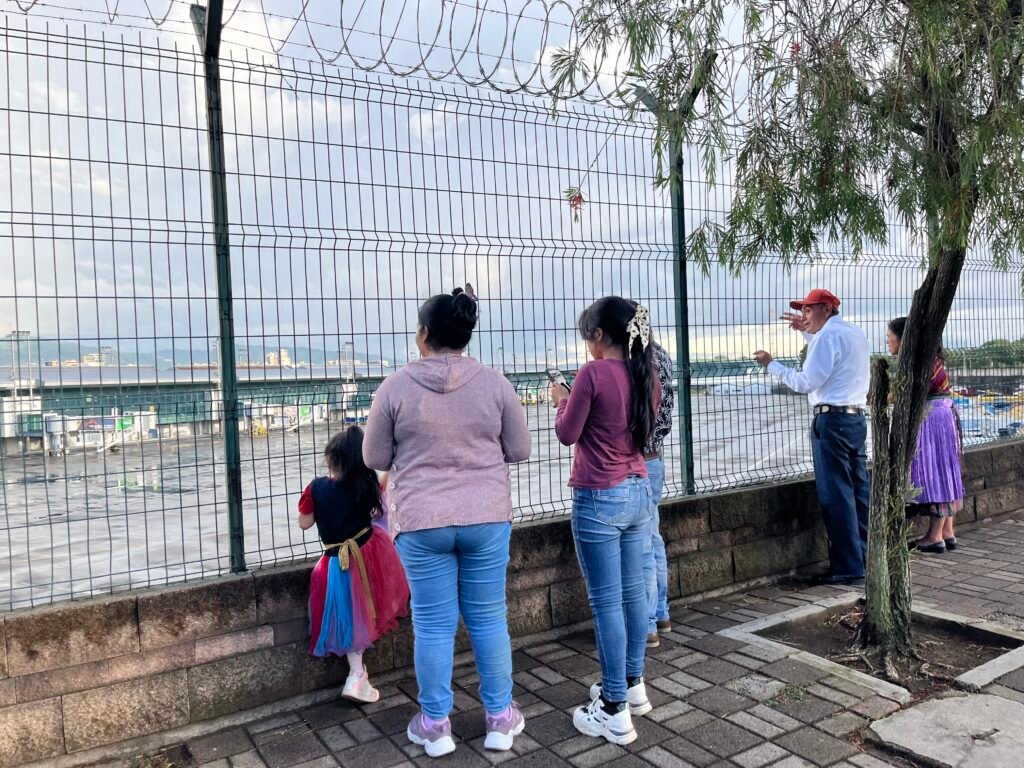 Familia, una niña, una señora, una joven, un señor, un anciano y una señora, mirando el aeropuerto desde la reja.