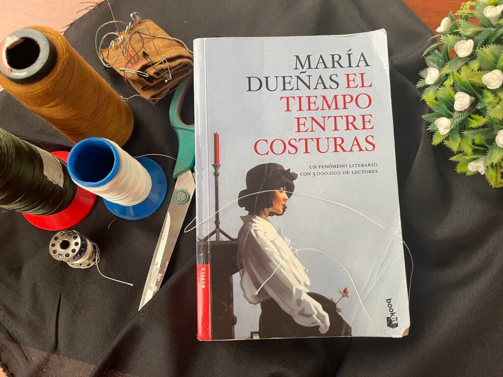 Carátula del libro El tiempo entre costuras de María Dueñas junto a hilos, tijeras, agujas, carretes y flores