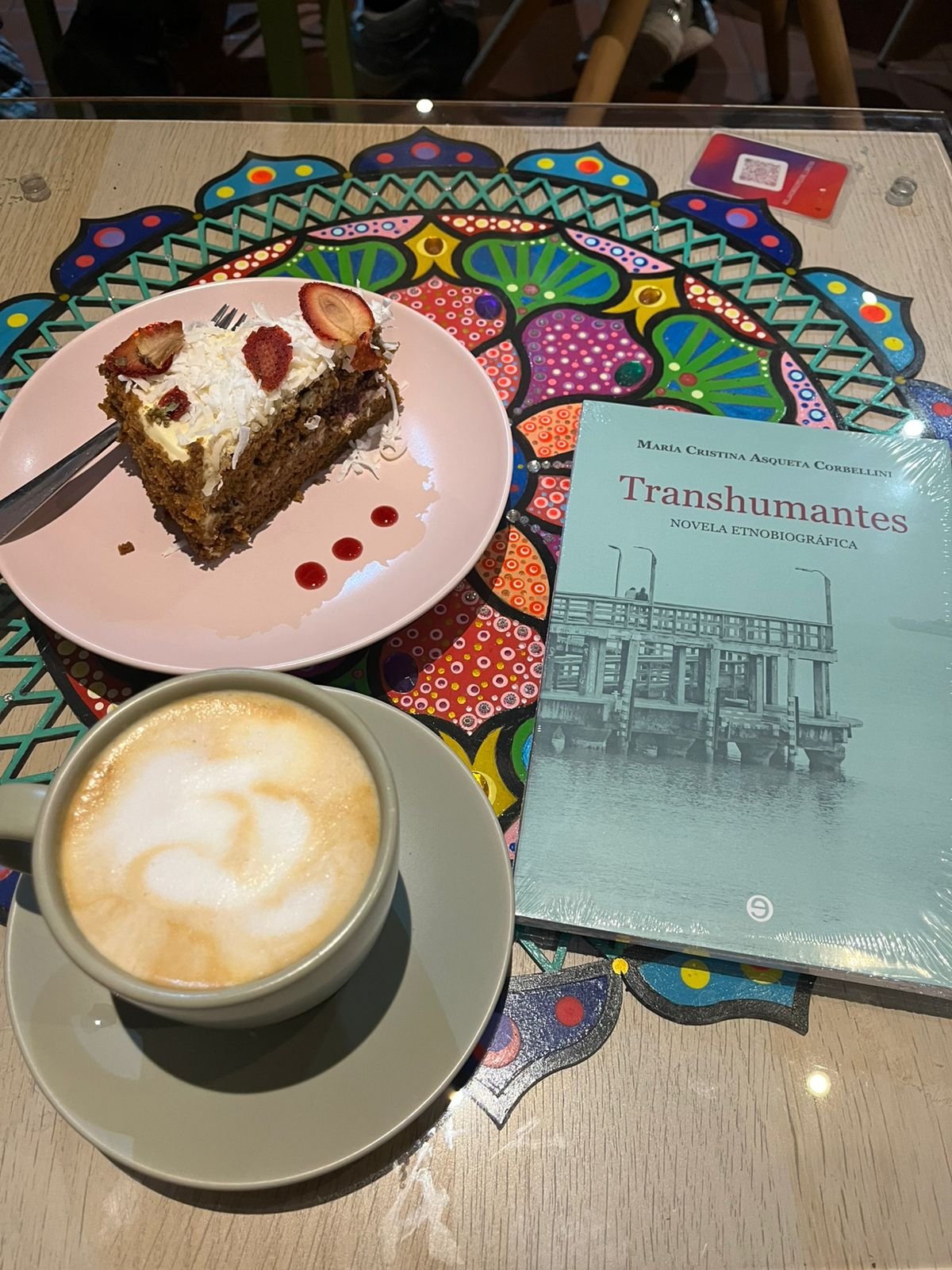 Torta de fresas secas con coco, un café caliente y el libro Transhumantes sobre una mesa de colores