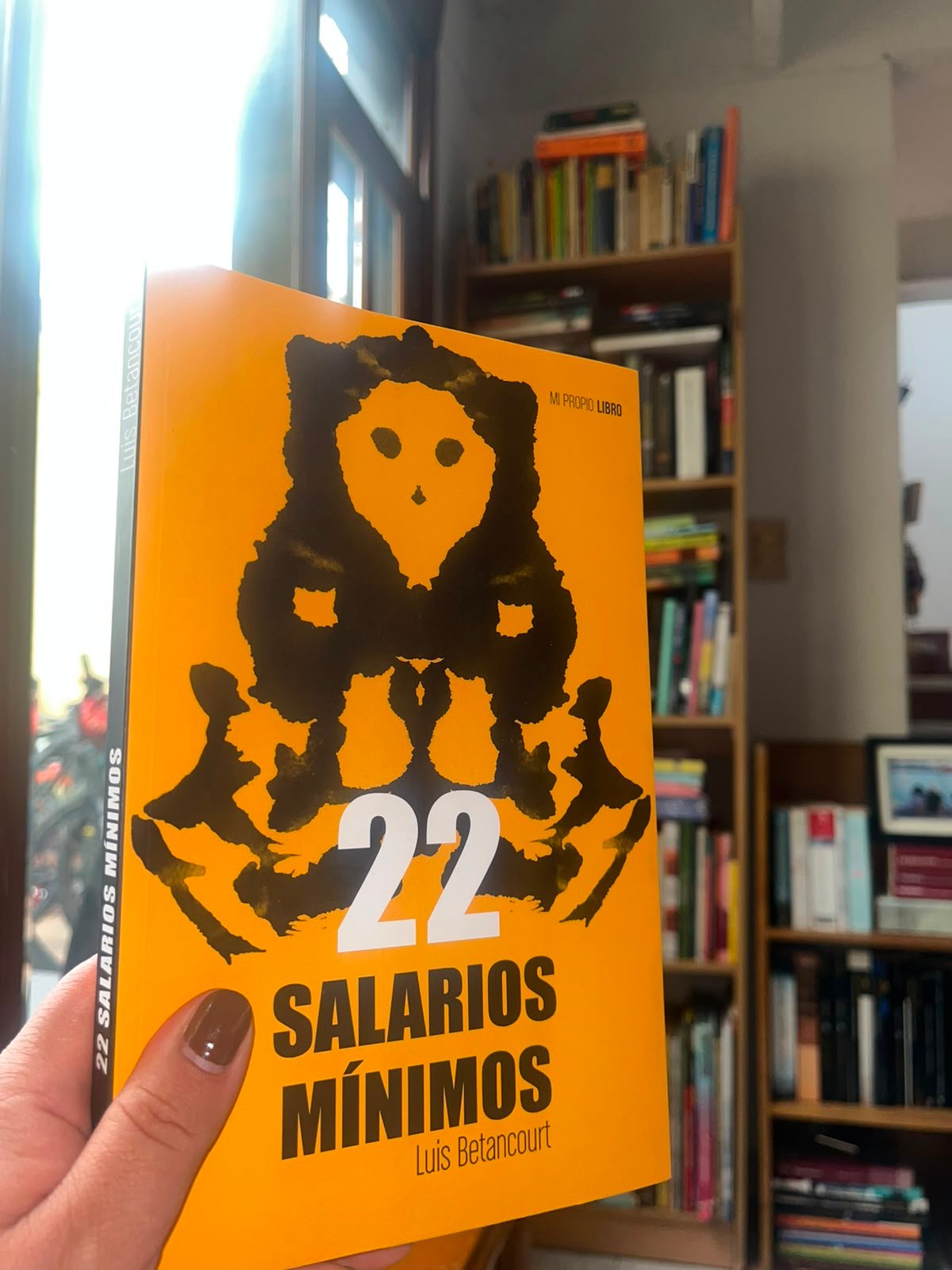 Portada del libro 22 salarios mínimos de Luis Betancourt, al fondo la biblioteca de Diana Socha
