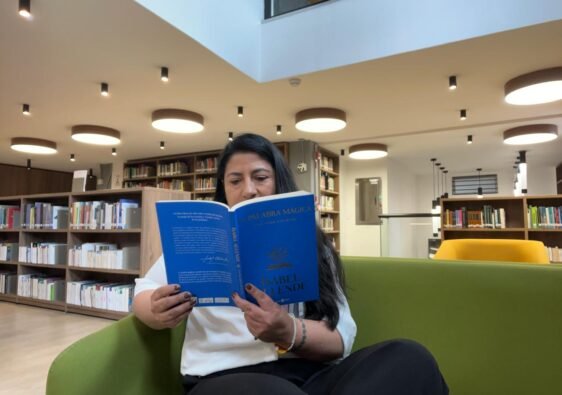 Diana en una biblioteca, al fondo se ven libros y ella está sentada en un sofá verde leyendo la palabra mágica de Isabel Allende