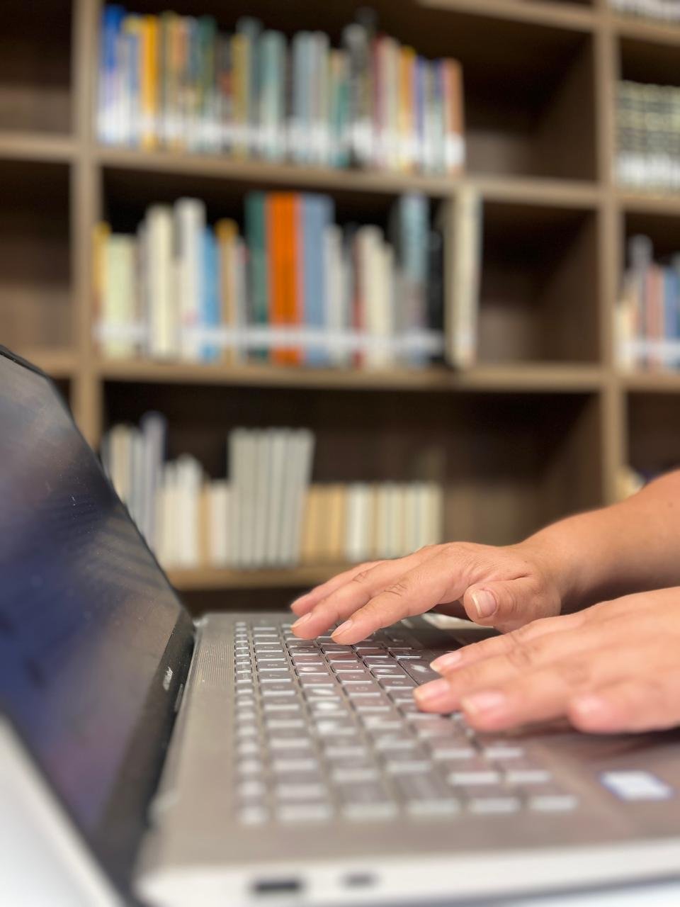 Al fondo se ve una biblioteca y en primer plano están las manos sobre un teclado del computador, simulando escribir