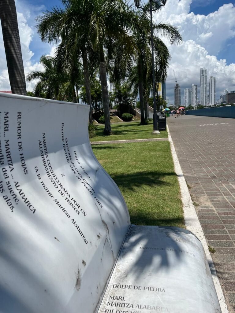Una silla en forma de libro. Se lee un poema de una escritora. Al fondo está una palmera y muy al fondo la ciudad de Panamá